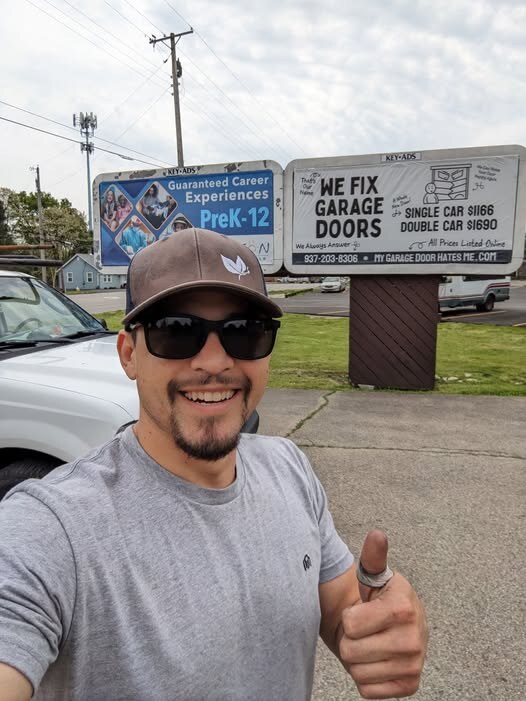 We Fix Garage Doors owner in Dayton, prices posted on the billboard behind him